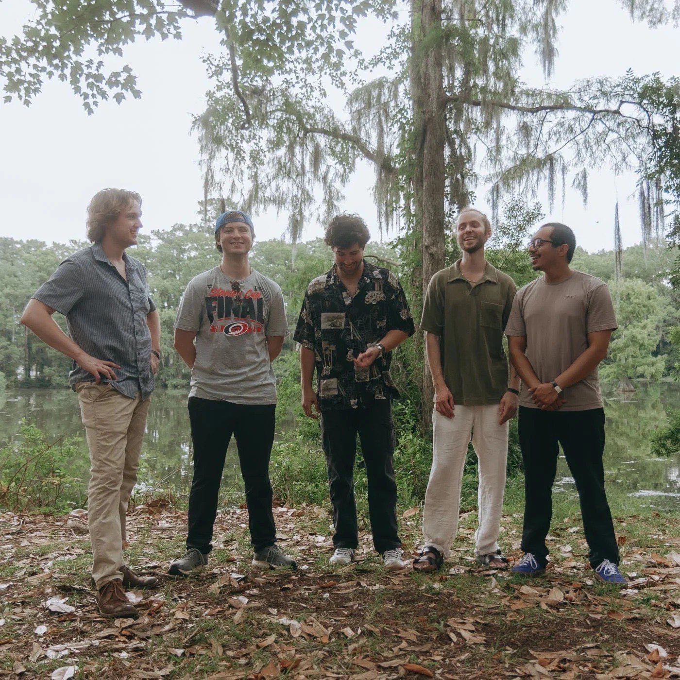 The five members of Jacoozy standing in a row under moss-hung trees at the water’s edge